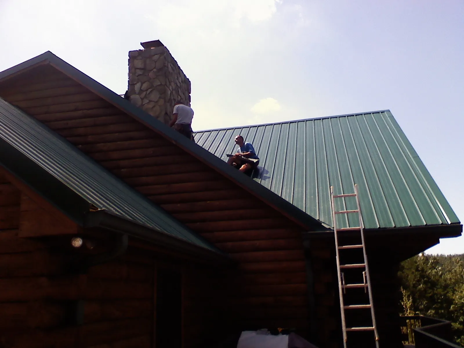 Roofers installing green metal panels beside a stone chimney on a log home in Diberville